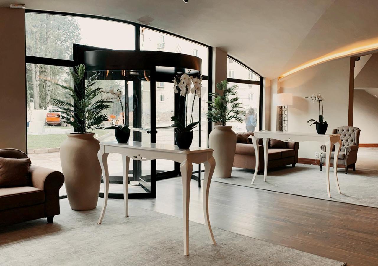 Buona Vitta Gramado Resort & Spa By Gramado Parks Экстерьер фото The photo displays an elegant interior space, likely a hotel or a high-end lobby. In the foreground, there is a stylish table with a curved design, positioned near large glass doors leading outside. Flanking the table are two large planters with lush