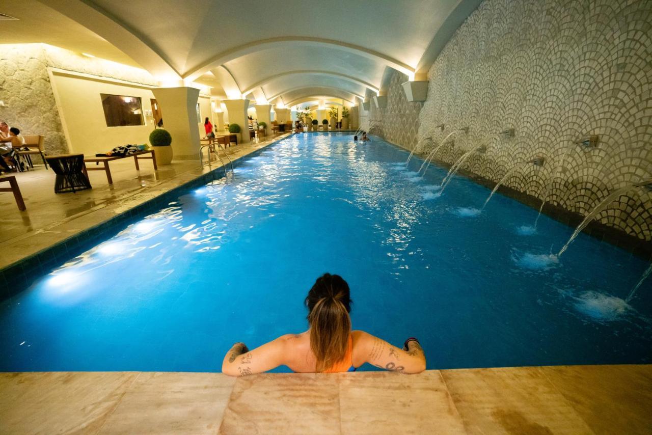 Buona Vitta Gramado Resort & Spa By Gramado Parks Экстерьер фото The photo shows an indoor swimming pool with a long, narrow design. The water is a clear blue, and there are features such as fountains along one side. In the foreground, a person with long hair is sitting on the edge of the pool, looking out over th