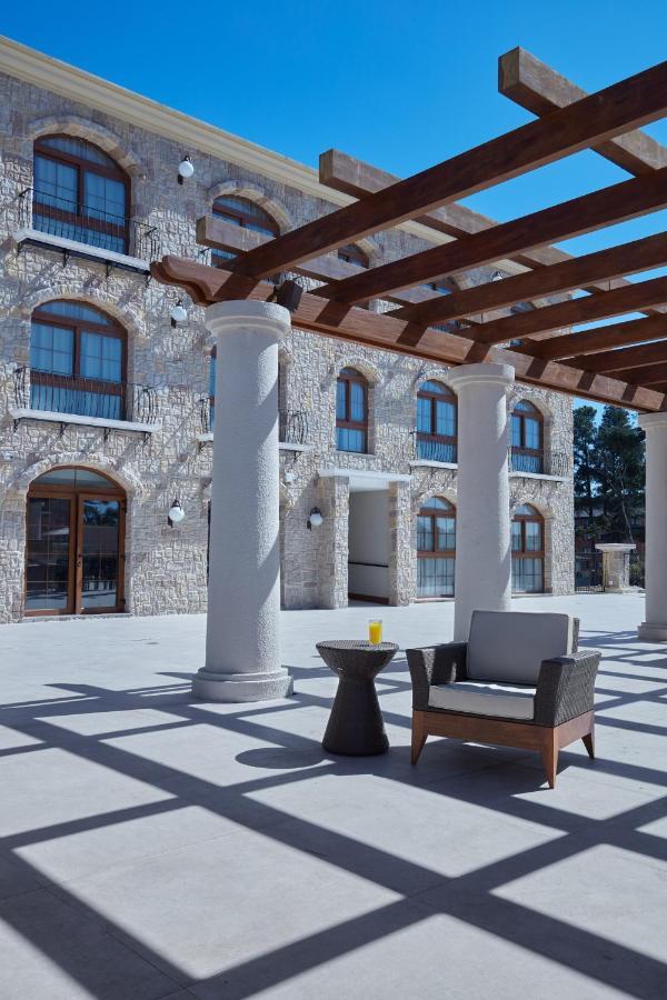 Buona Vitta Gramado Resort & Spa By Gramado Parks Экстерьер фото The photo shows a well-designed outdoor space with a stone wall building in the background. There are several tall columns that support a wooden pergola overhead, creating a shaded area. In this space, there is a stylish chair and a small table besid