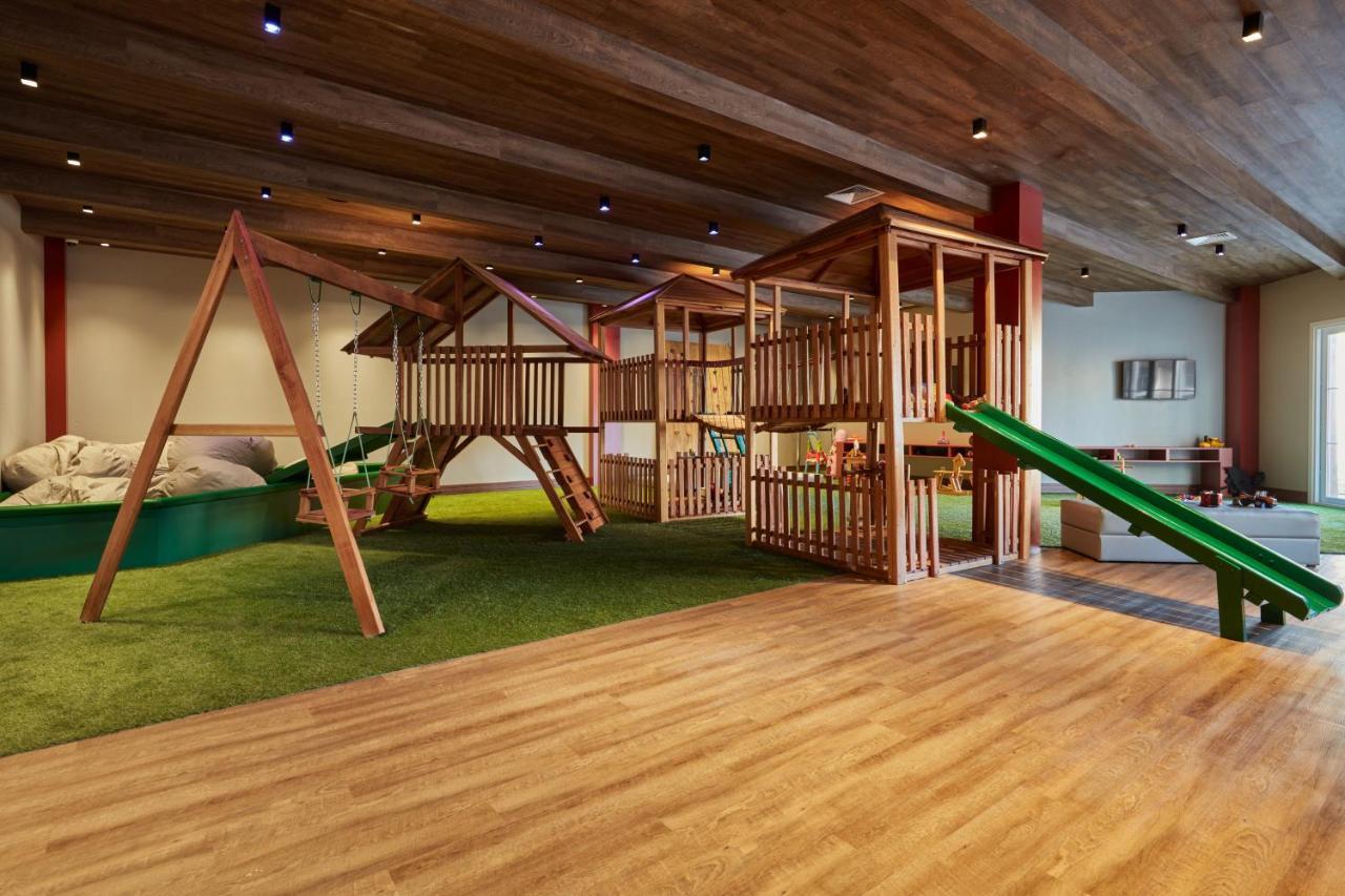 Buona Vitta Gramado Resort & Spa By Gramado Parks Экстерьер фото The photo shows an indoor play area designed for children. It features a wooden play structure that includes multiple levels, slides, and a swing set. The floor is covered with wooden planks, and there is a green carpet-like area that adds a soft tou