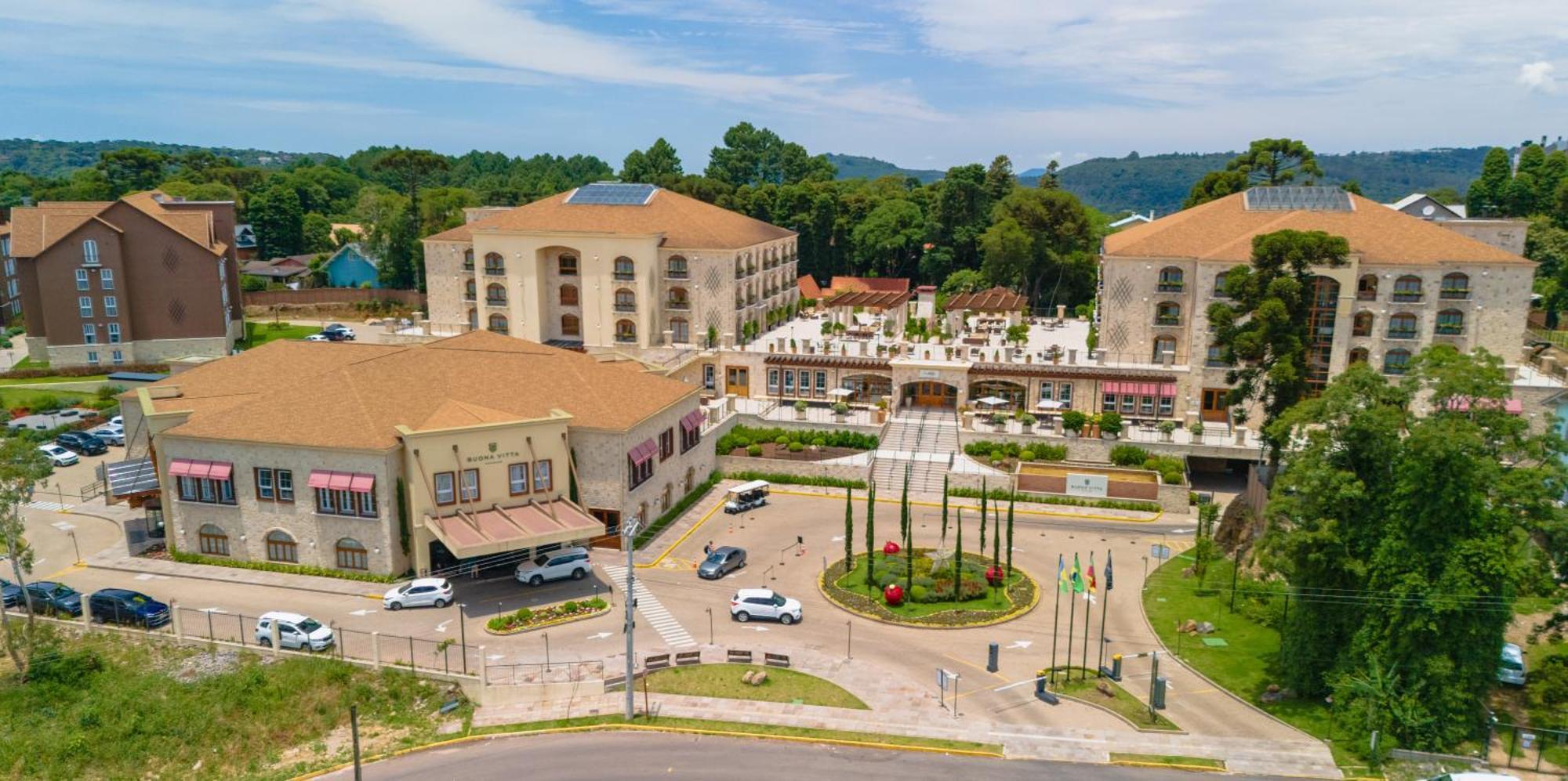 Buona Vitta Gramado Resort & Spa By Gramado Parks Экстерьер фото The photo shows an aerial view of a hotel complex surrounded by lush greenery. The buildings are designed with beige and light-colored facades, featuring multiple stories and a spacious layout. In the foreground, there is a circular road with several