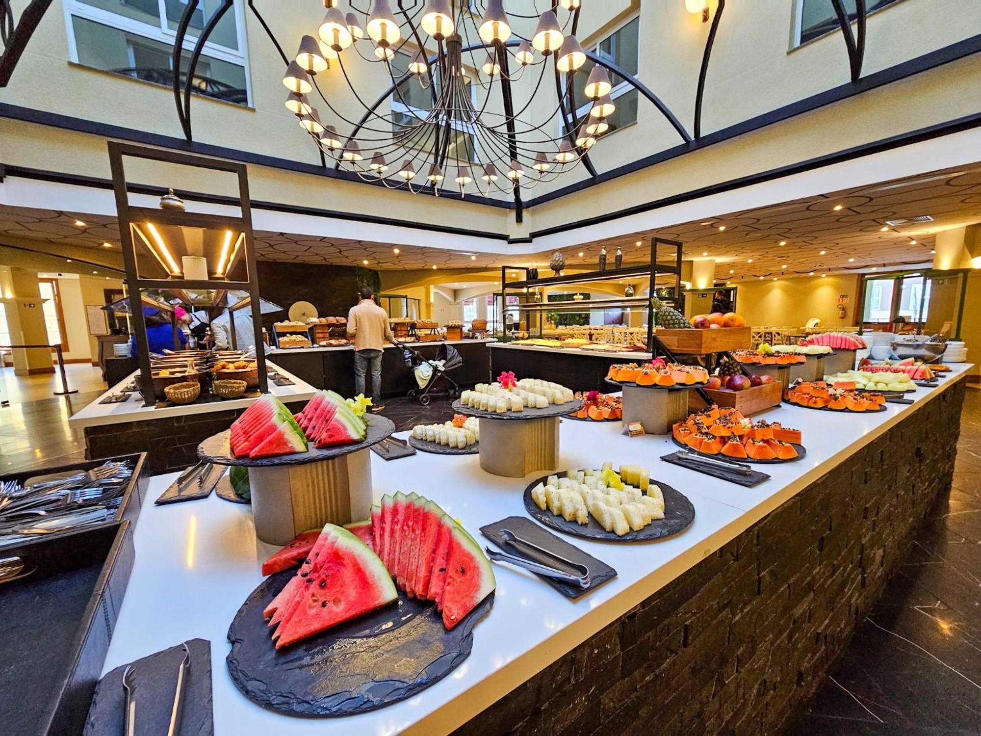 Buona Vitta Gramado Resort & Spa By Gramado Parks Экстерьер фото The photo depicts a lavish buffet setup in a dining area. In the foreground, there are several platters featuring an assortment of colorful food items, primarily fruits like watermelon and various types of sushi or rolls. The buffet table is elegantl