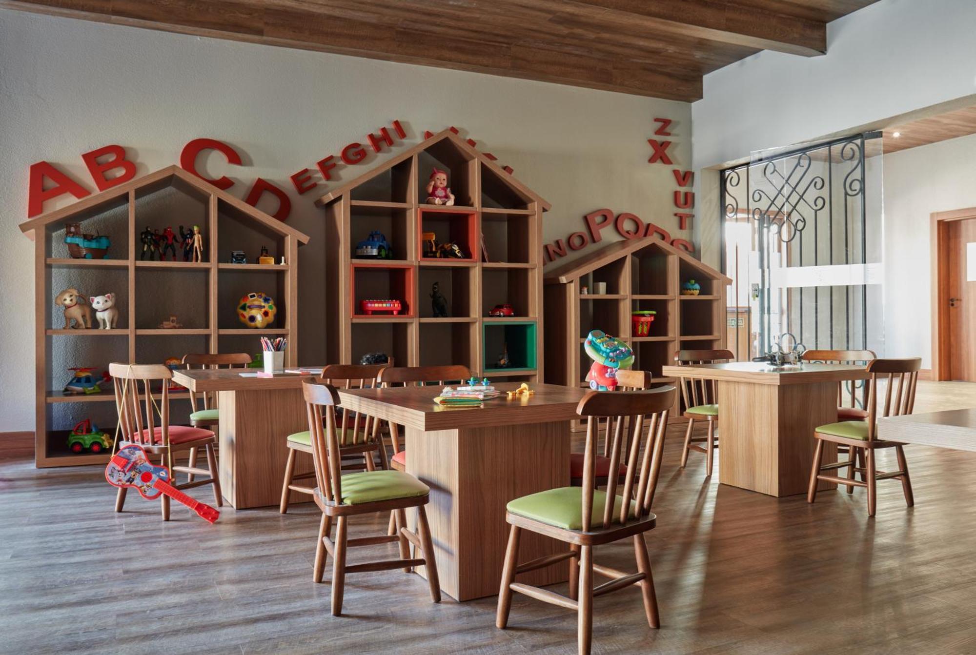 Buona Vitta Gramado Resort & Spa By Gramado Parks Экстерьер фото The photo shows a classroom or playroom designed for children. It features wooden furniture, including several tables and chairs with green cushions. In the background, there are large, open shelving units shaped like houses, displaying various toys 
