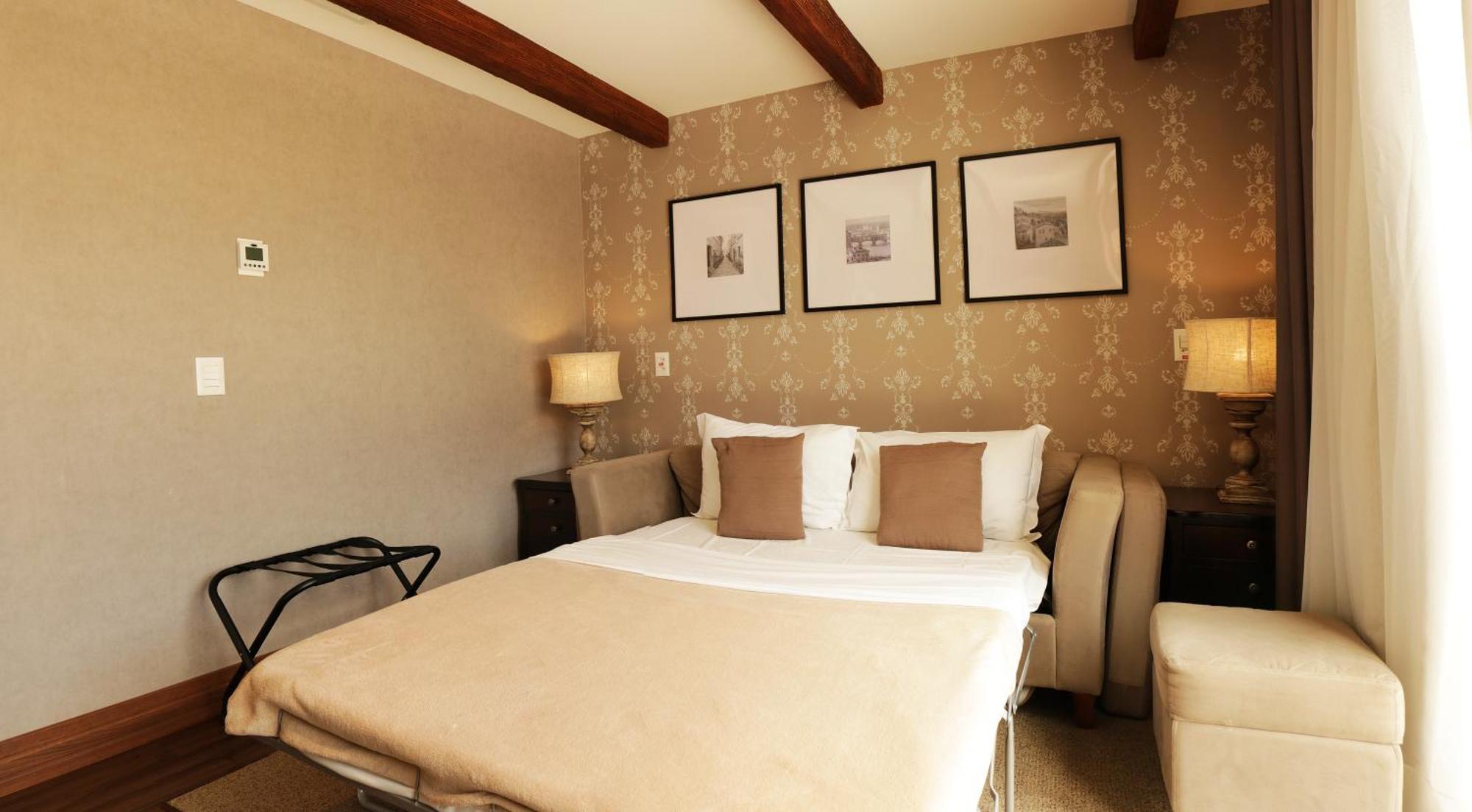 Buona Vitta Gramado Resort & Spa By Gramado Parks Экстерьер фото The photo shows a cozy bedroom with a neutral color palette. There is a bed with white linens and two brown decorative pillows. On either side of the bed, there are bedside tables with lamps. The walls feature a subtle floral pattern, and three frame