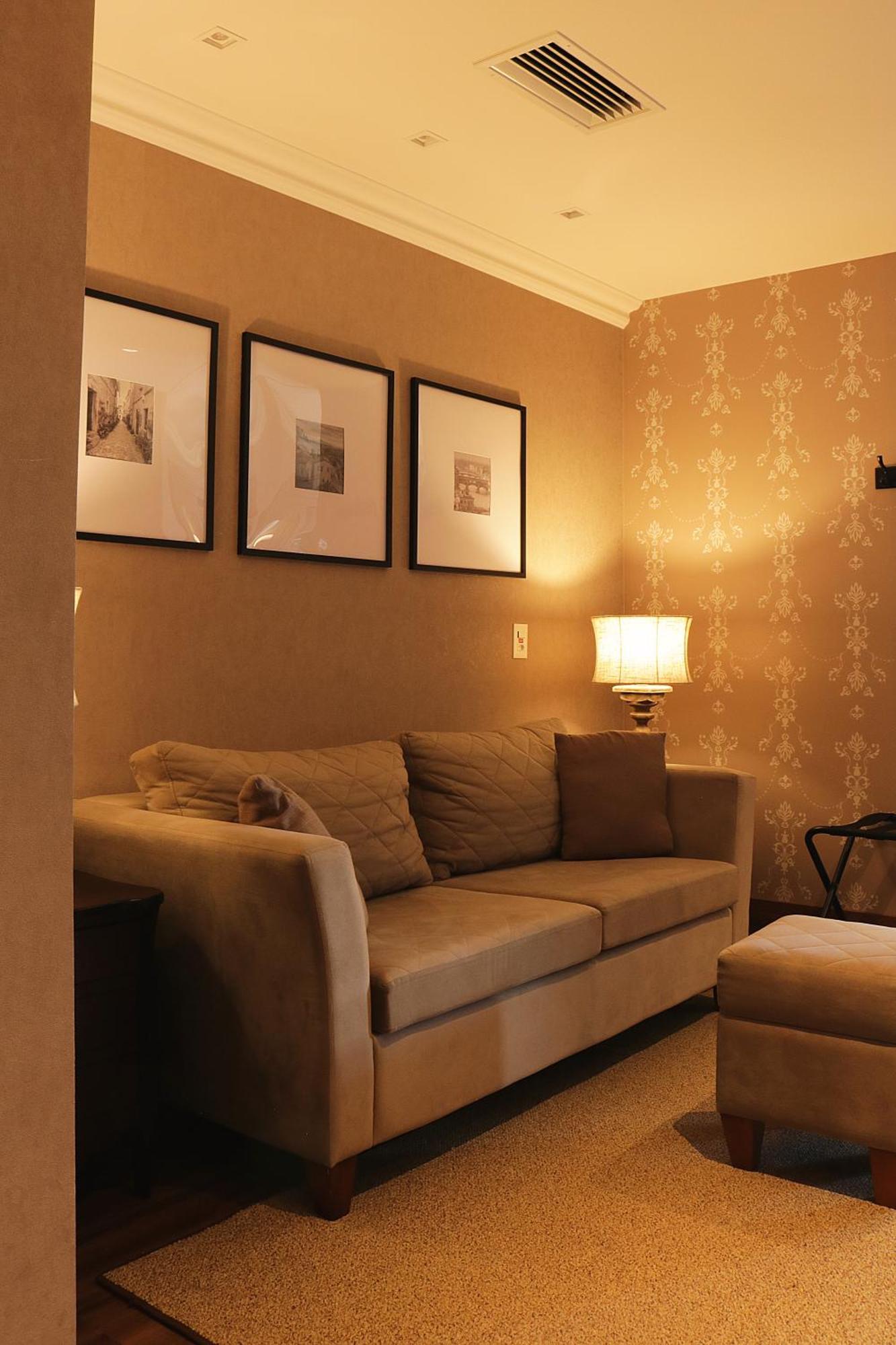 Buona Vitta Gramado Resort & Spa By Gramado Parks Экстерьер фото The photo shows a cozy living room setting. There is a light-colored sofa adorned with several cushions, positioned against a wall that has a subtle pattern. Above the sofa, there are three framed artworks or photographs hanging on the wall. To the s