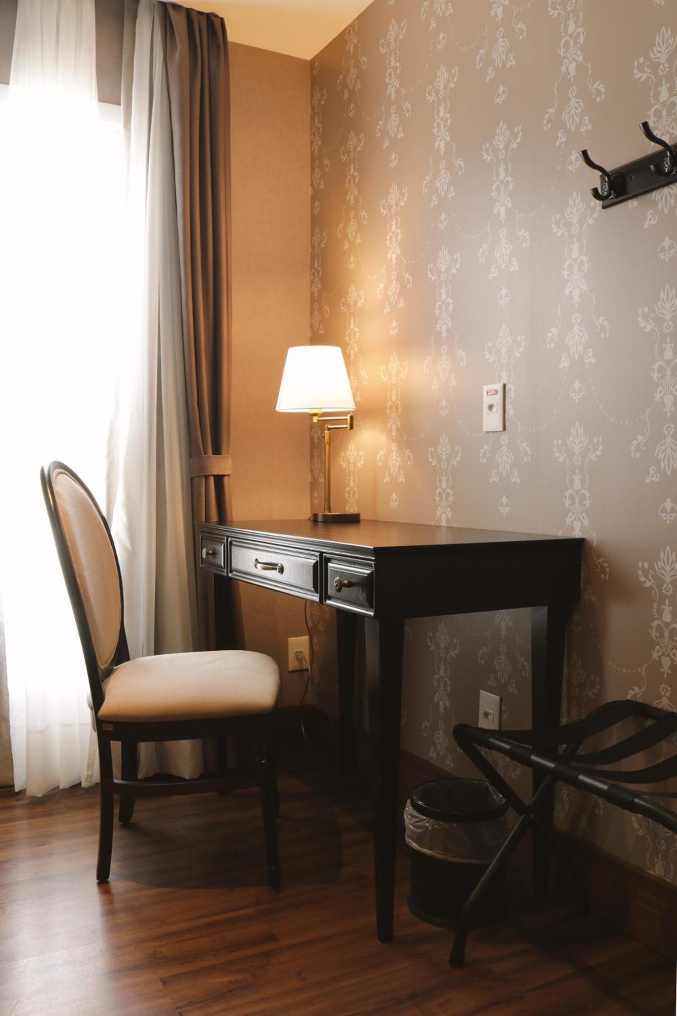 Buona Vitta Gramado Resort & Spa By Gramado Parks Экстерьер фото The photo shows a small desk set against a wall with a decorative pattern. The desk is black with a few drawers and has a small lamp on it. Next to the desk, there is a chair with a light-colored cushion. Soft light is coming through a window, castin