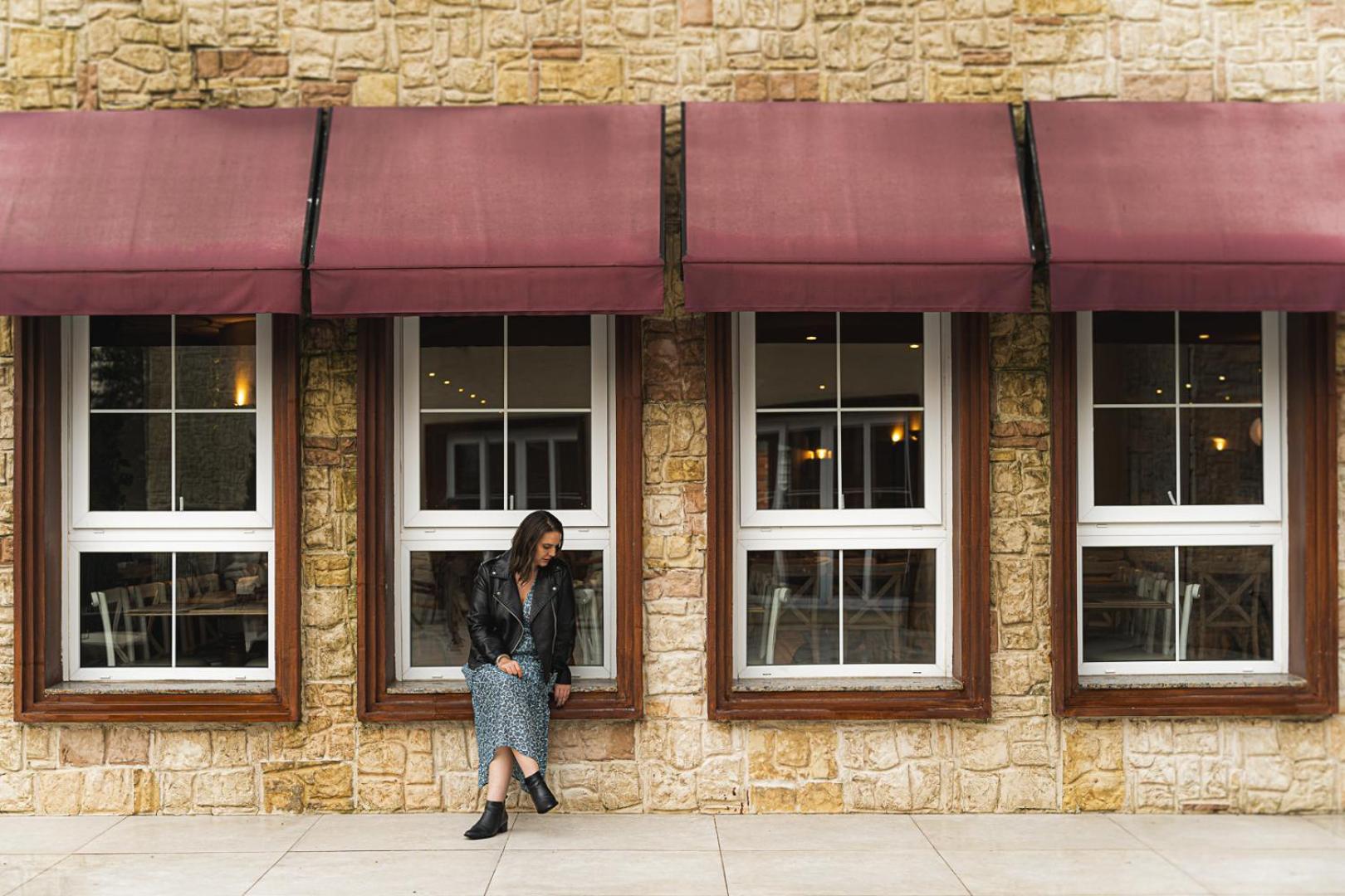 Buona Vitta Gramado Resort & Spa By Gramado Parks Экстерьер фото The photo shows a woman standing against a stone wall with three large windows in front of her, each framed with wooden borders. The windows have maroon awnings above them. The woman is dressed in a long skirt and leather jacket, leaning slightly aga